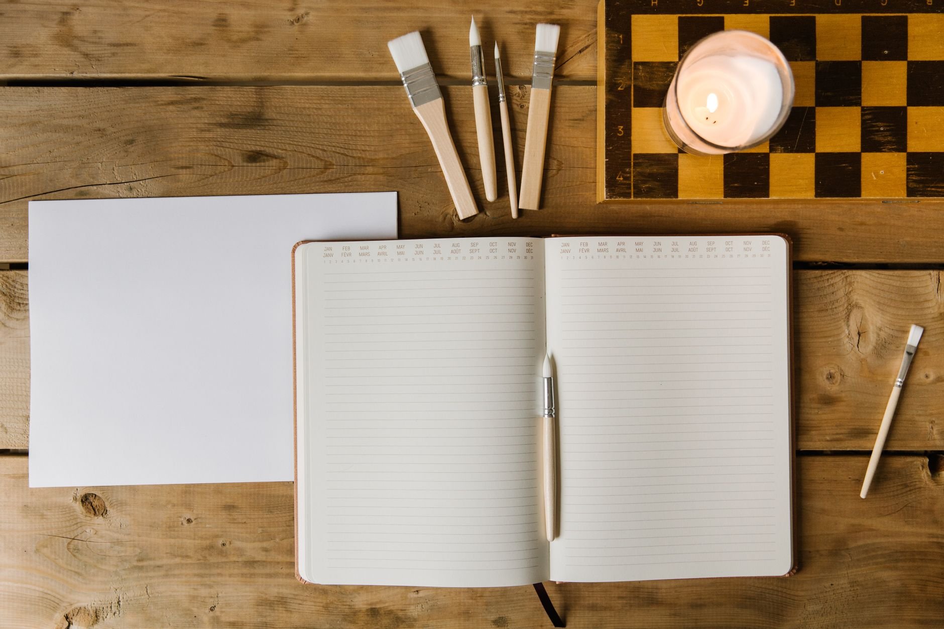 Open notebook with paintbrushes and a lit candle on a rustic wooden desk — analog warmth meets digital clarity
